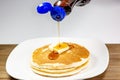 Pouring syrup from a blue tip bottle onto the square of butter on a stack of golden pancakes waiting to be eaten