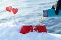 Pouring tea from a thermos in the on a picnic on Valentine`s Day. Red mugs with hot tea, lollipops hearts in the snow