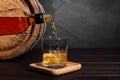 Pouring whiskey from bottle into glass with ice cubes near wooden barrel on table, space for text Royalty Free Stock Photo