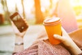 POV pretty woman lying on the bench in spring park and holding mobile phone and orange cardboard cup of tea or coffee. Royalty Free Stock Photo