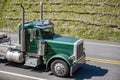 Classic bonnet day cab green big rig semi truck with high exhaust pipes transporting flat bed semi trailer running on the road Royalty Free Stock Photo