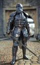 Powerful medieval knight standing with a full suit of armor and holding a sword weapon in front of his castle.