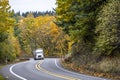 Powerful white big rig semi truck with covered bulk semi trailer climbing up hill on the winding mountain road with autumn forest Royalty Free Stock Photo