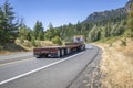 Powerful yellow big rig semi truck with empty flat bed semi trailer running on the winding road with trees and mountain to Royalty Free Stock Photo