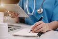 Practitioner in lab room writing on blank notebook and work on tablet with medical stethoscope on the desk at hospital.