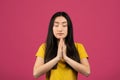 Pray and meditate. Portrait of chinese lady keeping palms together and praying, meditating, pink background, studio shot