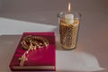 Prayer book with pink binding with wooden Christian Catholic rosary on it lying near lighted candle standing in glass among peas