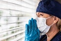 Prayerful Stressed Female Doctor or Nurse Crying At Window Wearing Medical Face Mask