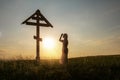 praying woman in front wooden Cross on a hilltop at sunset.