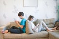 Pre-teen sister and brother gaming online with cellphones while sitting at home, children wearing safety masks, sitting back to