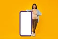 Pregnancy app. Happy expectant lady standing near huge smartphone with blank white screen, mockup