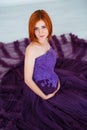Pregnancy beauty. Beautiful elegant readhead pregnant woman in violet dress posing in tender home interior.
