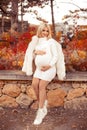 Pregnancy. Motherhood. Pregnant woman hugging her belly enjoying in autumn forest park. Beautiful blond woman in white knit