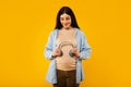 Pregnancy and music. Happy expectant lady holding headphones near belly and smiling, yellow studio background