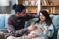 Pregnancy and people concept - happy African man hugging pregnant woman at home. Young couple sitting on sofa, husband support and Royalty Free Stock Photo