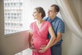 Pregnancy and people concept - happy man hugging his pregnant wife standing at window Royalty Free Stock Photo