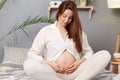 Pregnancy, rest, people and expectation. pregnant woman with bare belly wearing white clothing smiling sitting in bed and touching