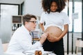 Pregnant african woman has appointment with doctor at clinic. Male gynaecologist OB GYN medic specialist with stethoscope listens