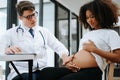 Pregnant african woman has appointment with doctor at clinic. Male gynaecologist OB GYN medic specialist with stethoscope listens Royalty Free Stock Photo