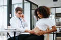 Pregnant african woman has appointment with doctor at clinic. Male gynaecologist OB GYN medic specialist with stethoscope listens