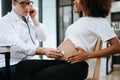 Pregnant african woman has appointment with doctor at clinic. Male gynaecologist OB GYN medic specialist with stethoscope listens