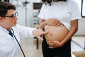 Pregnant african woman has appointment with doctor at clinic. Male gynaecologist OB GYN medic specialist with stethoscope listens