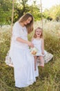 Pregnant beautiful mother with little blonde girl in a white dress sitting on a swing, laughing, childhood, relaxation Royalty Free Stock Photo