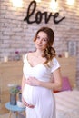 Pregnant beautiful woman wearing white dress holding belly, inscription love on brick wall.