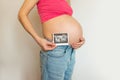 Pregnant belly girl with ultrasound in her hands on a light background. Smiling young pregnant in jeans woman hugs her belly with
