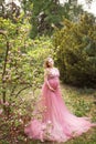Pregnant blonde in long pink fattini dress touches belly and looks at sky
