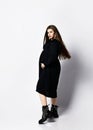 Pregnant woman in long black dress and brutal shoes stands side to camera holding hands on her belly and looks at camera Royalty Free Stock Photo