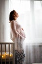 A pregnant Caucasian woman in a knitted shawl stands leaning on a baby`s cradle and hugs her tummy. The window in the background. Royalty Free Stock Photo