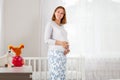 A pregnant Caucasian woman stands in the children`s white room and hugs her tummy. In the background, a large window and a cradle Royalty Free Stock Photo