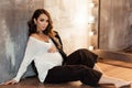 Pregnant european woman in white blouse and black pants is sitting near the wall in a dark flat, stylish and happy