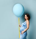 Pregnant female in blue dress. Smiling, put hand on tummy, holding balloon by yellow ribbon, posing sideways on blue background Royalty Free Stock Photo