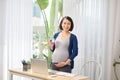 Pregnant freelancer touching belly near laptop, glass of water on table