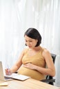 Pregnant freelancer touching belly near laptop, writing on diary on table