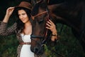 Pregnant girl with a big belly in a hat next to horses in the forest in nature.Stylish girl in white clothes and a brown jacket Royalty Free Stock Photo