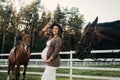 A pregnant girl with a big belly in a hat next to horses near a paddock in nature.Stylish pregnant woman in a brown dress with Royalty Free Stock Photo