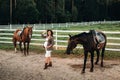 A pregnant girl with a big belly in a hat next to horses near a paddock in nature.Stylish pregnant woman in a brown dress with Royalty Free Stock Photo