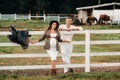 A pregnant girl in a hat and her husband in white clothes stand next to the horse corral.a stylish couple waiting for a child Royalty Free Stock Photo