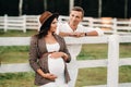 A pregnant girl in a hat and her husband in white clothes stand next to the horse corral.a stylish couple waiting for a child Royalty Free Stock Photo