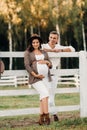 A pregnant girl in a hat and her husband in white clothes stand next to the horse corral.a stylish couple waiting for a child Royalty Free Stock Photo