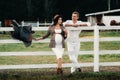 A pregnant girl in a hat and her husband in white clothes stand next to the horse corral.a stylish couple waiting for a child Royalty Free Stock Photo