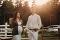 A pregnant girl in a hat and her husband in white clothes stand next to the horse corral.a stylish couple waiting for a child Royalty Free Stock Photo