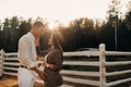 A pregnant girl in a hat and her husband in white clothes stand next to the horse corral.a stylish couple waiting for a child Royalty Free Stock Photo