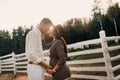 A pregnant girl in a hat and her husband in white clothes stand next to the horse corral.a stylish couple waiting for a child Royalty Free Stock Photo