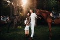 A pregnant girl in a hat and her husband in white clothes stand next to horses in the forest in nature.Stylish pregnant woman with Royalty Free Stock Photo