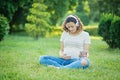 Happy young pregnant woman sitting on carpet and listening music on headphones from smartphone at park. Pregancy, Relaxation and Royalty Free Stock Photo