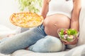 Pregnant girl sitting on a sofa holding salad bowl in one hand and pizza in the other one Royalty Free Stock Photo